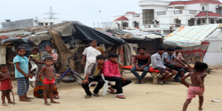 A slum with its residents in front of it and a big house behind to indicate wealth inequality